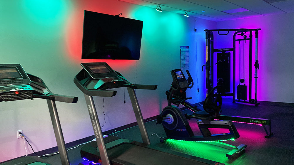 Treadmill, stationary bike, row machine, cable machine, and TV inside a gym with colorful lighting.